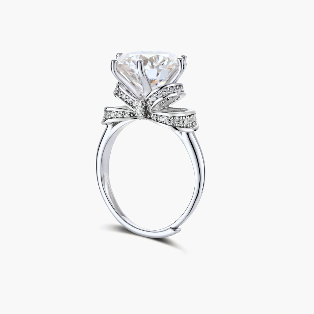 Sterling silver ring with 5ct round moissanite center stone and intricate bow design with pavé accents on white background.